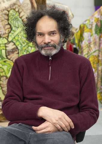 The artist Hew Locke wearing a purple top and sat in a chair.
