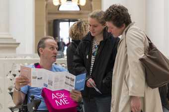a man in a wheelchair shows two visitors a map inside Tate Britain
